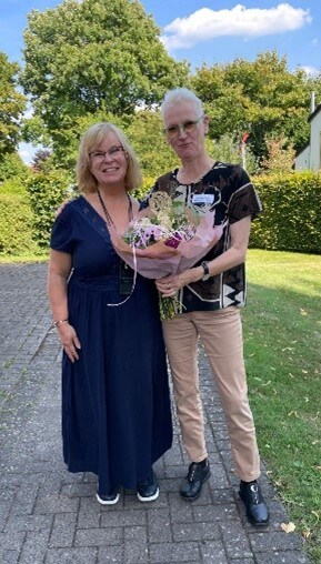 Zwei Frauen stehen auf einem gepflasterten Weg. Eine trägt ein dunkelblaues Kleid, die andere eine schwarze Bluse mit Muster und beige Hose. Beide halten einen großen Blumenstrauß.