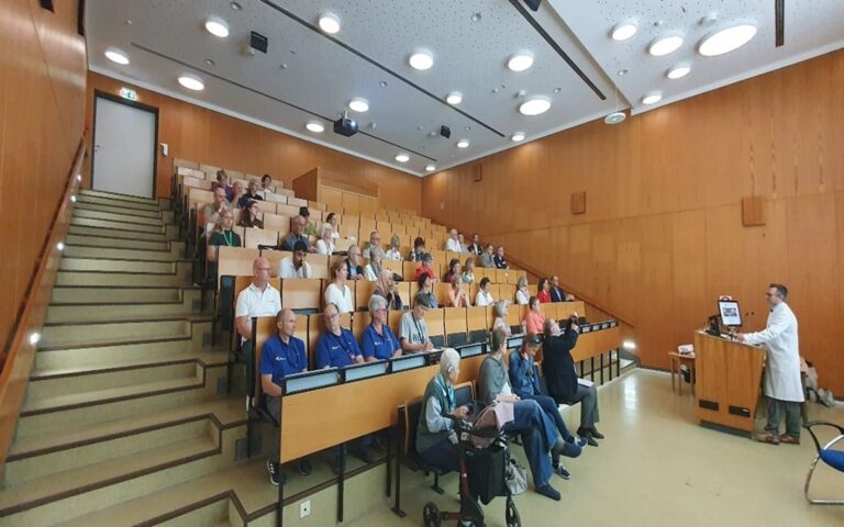 Ein Hörsaal mit mehreren Reihen von Holzstühlen, besetzt von etwa 30 Personen. Ein Referent steht an einem Tisch vorne und präsentiert