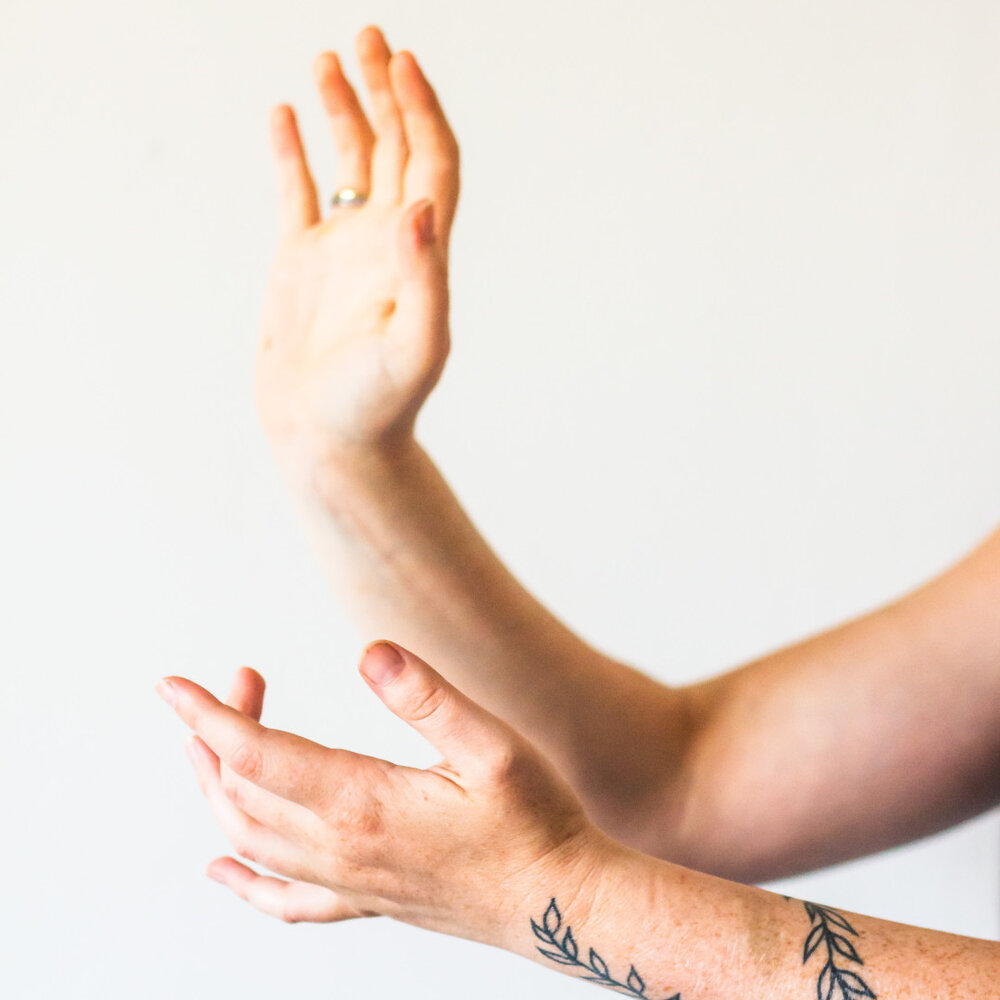 Zwei ausgestreckte Arme mit sichtbaren Tätowierungen auf dem Unterarm, eine Hand zeigt nach oben, die andere nach vorne.